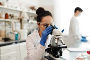 Woman in laboratory wearing lap coat and gloves looking through microscope